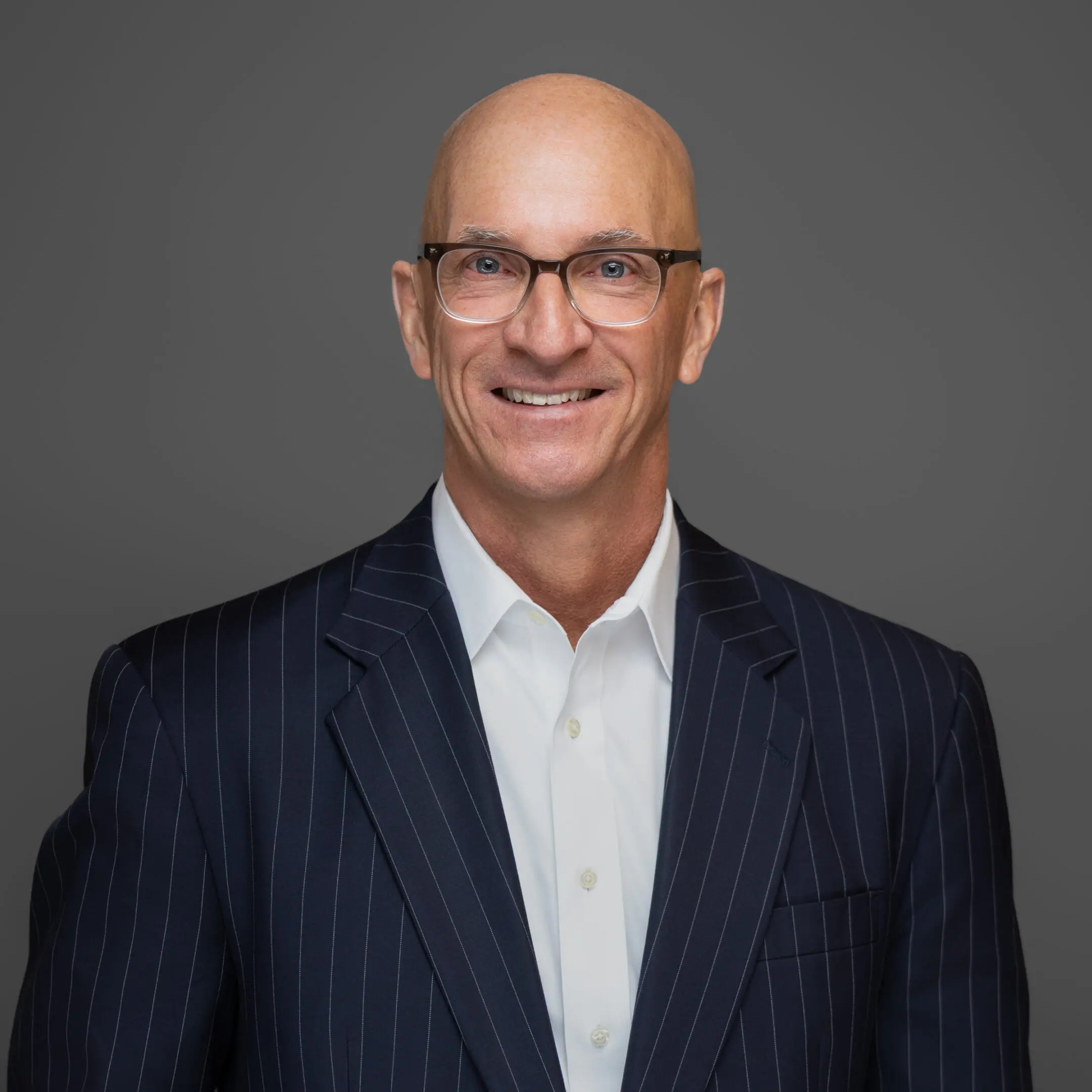 Smiling bald man in glasses wearing a dark pinstripe suit and white shirt.