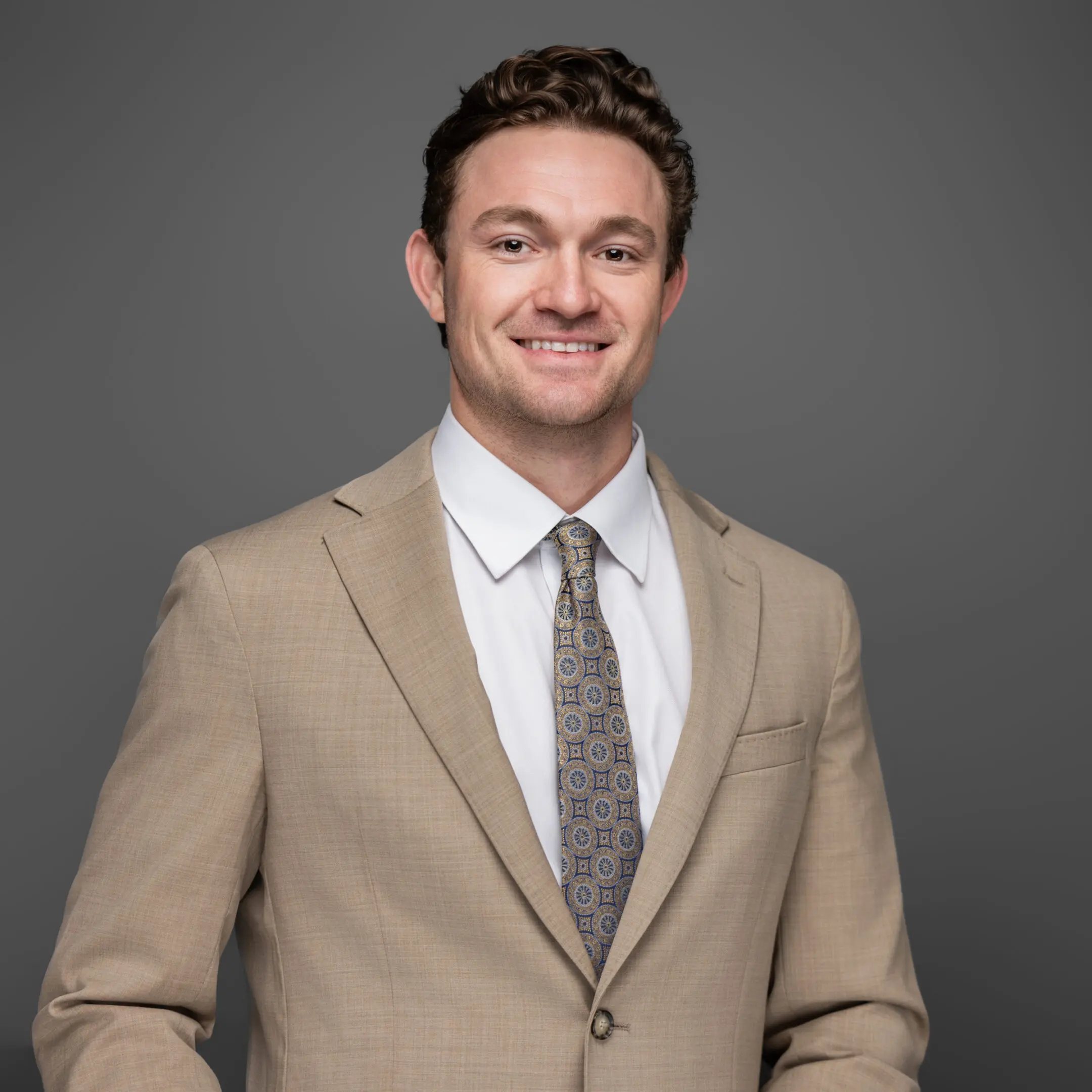 Smiling man in beige suit and patterned tie against gray background.