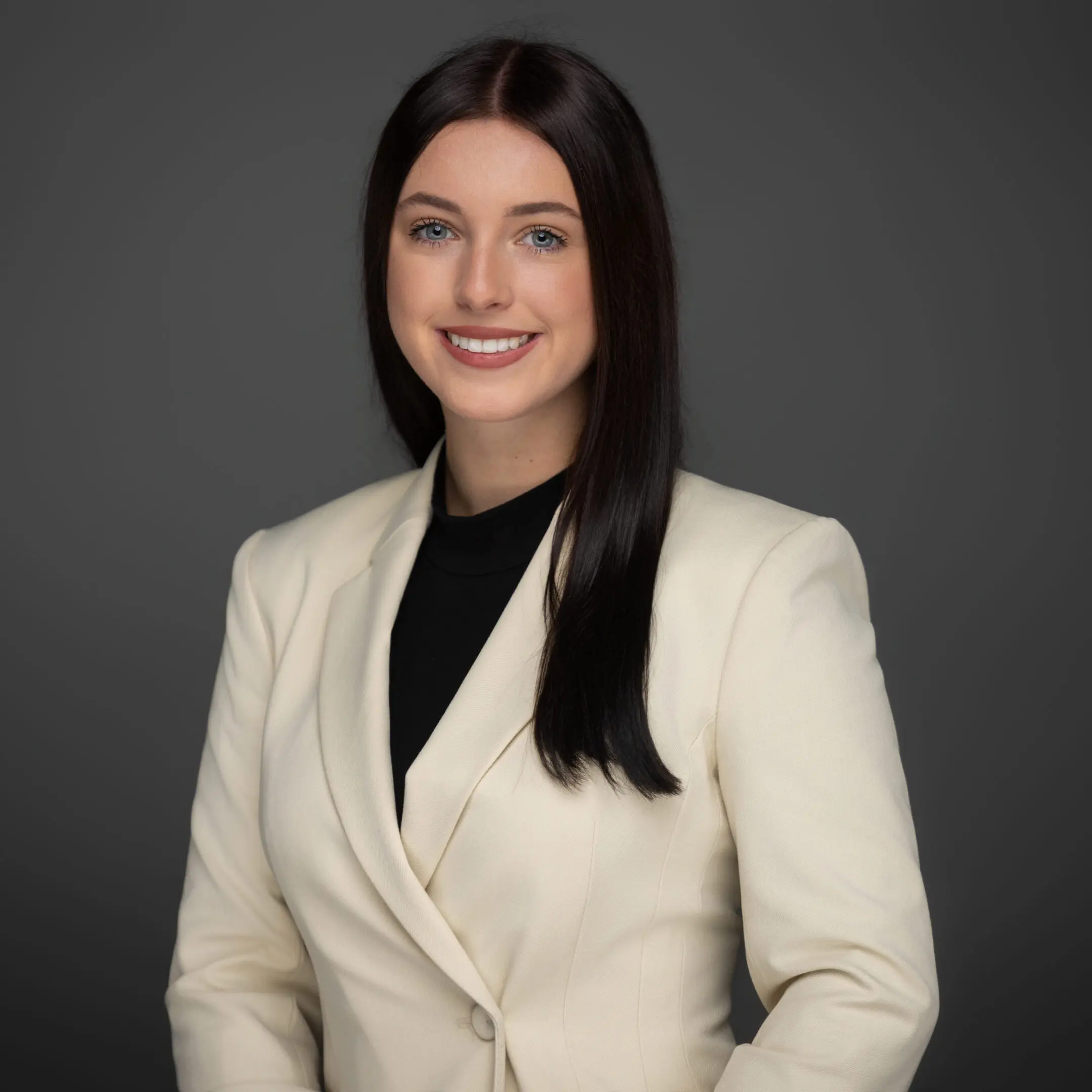 A woman with long dark hair in a cream blazer and black top smiles at the camera against a plain gray background, ideal for corporate headshots.